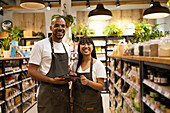 Cheerful young African American salesmen in aprons standing between cupboards with grocery items holding a tablet while smiling looking at camera