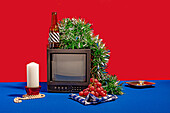 Vintage-Fernsehgerät, umgeben von einer Reihe von Objekten, darunter eine Flasche mit gestreiftem Etikett, frische Trauben auf einem karierten Tuch, eine weiße Kerze und grünes Lametta, alles vor einem roten Hintergrund auf einem blauen Tisch mit einem Gertenfuß mit festlich verzierter Socke