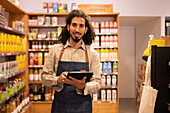 Portrait of happy ethnic male worker with long curly hair in casual clothes standing near shelves looking at camera smiling while working on tablet in modern lighted grocery shop