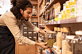 Side view of young ethnic bearded male seller with curly hair in denim apron standing in supermarket with glass jar of bulk food during work