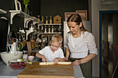 Mid adult Caucasian mother dressed in casuals looking at cute little son cutting banana into slices with knife on chopping board in kitchen