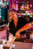 Focused beautiful caucasian young barmaid pouring drink in mug from glass bottle at illuminated counter in modern bar
