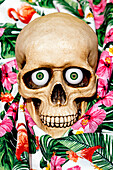Gruseliger menschlicher Schädel, der mit grünen Augen in die Kamera blickt, während er auf Stoff mit gezeichneten Blumen gelegt wird