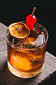 Hochformatiges Glas, gefüllt mit alkoholischem Old Fashioned Whiskey und Eiswürfeln, garniert mit getrockneten Zitronenscheiben und Kirschen auf dem Tisch