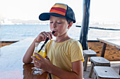 Cute thirsty boy in casual clothes and cap drinking pineapple juice in glass with straw while sitting in cafe against beach during summer vacation