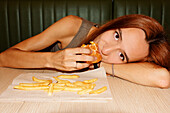Young woman with red hair and piercing looking at camera while laying on table and enjoy eating hamburger in restaurant