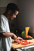 Side view of concentrated young ethnic African American African American male in white t shirt slicing ripe red bell pepper with sharp knife on wooden chopping board while preparing healthy salad in kitchen