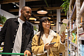 Low angle of cheerful multiethnic couple in casual clothes with shopping trolley and smartphone choosing goods while buying products together