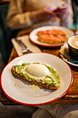 Leckerer Toast mit pochiertem Ei und zerdrückter Avocado auf Keramikteller neben einer Tasse Cappuccino in einem Cafe