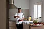 Positive young ethnic African American male in casual clothes preparing delicious dish while frying tomatoes on pan and mixing ingredients with wooden spatula during cooking process in kitchen