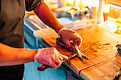From above of crop anonymous male chef wearing gloves and black uniform slicing fish with knife on illuminated wooden counter at modern sushi bar