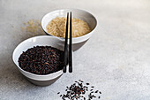 High angle of Raw wild black rice and peeled white rice in the bowls with chopsticks against white surface