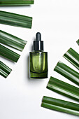 Green glass cosmetic bottle with pipete at white background with green palm leaves. Skin care with moisturizing face serum. Top view with copy space.
