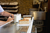 A glass of milk and a cake on a counter with a anonymous person arm in the background in a bakery setting