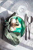 Top view of Easter table setting, showcasing a vibrant green ceramic plate with two decorative Easter eggs adorned with white and green patterns and delicate feathers, placed on gray surface between napkin and cutlery and glass of water