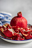 Closeup of ripe red pomegranate seeds on a white rustic plate with blue rim on a gray textured background.