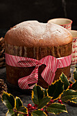 A traditional panettone dusted with icing sugar, adorned with a decorative ribbon, surrounded by holly leaves and berries, evoking the warmth of the holiday season