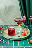 Glasses of refreshing red alcohol cocktails with strawberries and straw served on tray with berries and lemon slice on table