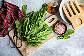 From above of vibrant spinach leaves placed on a wooden board, accompanied by a bowl of pink salt, a bottle of oil, wooden spatula, and rustic scissors on a textured backdrop