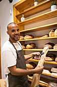Positive young African American male seller in apron putting fresh baked bread into paper bag while standing looking at camera near shelves working