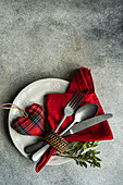 Draufsicht auf einen Keramikteller mit Besteck, eine Serviette aus Schottenstoff mit herzförmigem Dekor auf einer Betonfläche am Küchentisch für eine Mahlzeit während der Valentinstagsfeier