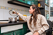 A woman in a wheelchair comfortably reaches for a pot lid while cooking in an adapted kitchen space