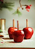 Rote Äpfel mit Zimtstangen auf Küchentisch mit Zucker an Wand Hintergrund mit Tannengrün. Mit Zucker glasierte Äpfel. Leckere Weihnachtssüßigkeiten. Vorderansicht