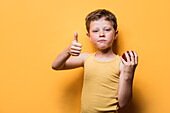 Portrait of cute boy in casual clothes showing thumbs up sign and holding red ripe apple while looking at camera and standing against yellow background
