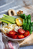 A healthy bowl of fresh vegetables featuring sliced cucumber, cherry tomatoes, cauliflower florets, a lemon wedge, and arugula leaves on a wooden board with a side of mixed nuts