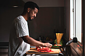 Side view of concentrated young ethnic African American African American male in white t shirt slicing ripe red bell pepper with sharp knife on wooden chopping board while preparing healthy salad in kitchen