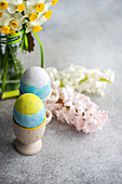 Osterkarte Konzept mit bunten Eiern auf Eierbechern in der Nähe von saisonalen Blumen auf konkreten Hintergrund