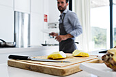 Side view of crop blurred male chef in apron looking at camera while standing near kitchen table with knife and cut pieces of pineapple on chopping board in daylight