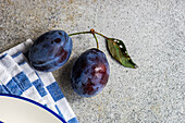 Closeup of ripe plums placed on gray surface near ceramic bowl and striped napkin