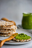 Rice bread on a plate accompanied by a vibrant green spinach pesto pasta-sauce in a glass jar, set against a blurred backdrop near salt shaker
