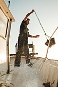 Bärtiger männlicher Fischer gießt Wasser aus einem Eimer auf dem schmutzigen Deck eines Schoners beim Fischen in Soller nahe der Baleareninsel Mallorca