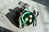 Top view of Easter table setting, showcasing a vibrant green ceramic plate with two decorative Easter eggs adorned with white and green patterns and delicate feathers, placed on gray surface between napkin and cutlery and glass of water