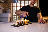 Anonymous bearded male chef in black shirt standing at table in kitchen and blowing gas flame on delicious uramaki sushi rolls while preparing to serve food against blurred interior