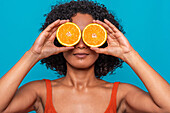 Serious young Hispanic female with curly hair covering eyes with slice of orange while standing against blue background