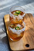 High angle of glasses with Japanese sochu liquor with mango juice and slices of Jalapeno pepper with salt on the glass edge placed on wooden table against blurred gray background