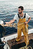 Von oben fokussierter bärtiger männlicher Fischer in Uniform, der mit einem Netz auf Fischfang geht, während er auf einem Schoner in Soller nahe der Baleareninsel Mallorca arbeitet