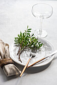 Hoher Winkel einer Tischdekoration mit einer frischen Pistazienpflanze auf einem Teller mit Besteck neben einer Serviette und einem Glas auf einer grauen Fläche im Tageslicht