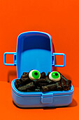 Horror lunch with black pasta and eyes in lunchbox placed on orange background