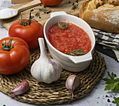 Von oben: reife Tomaten und ungeschälter Knoblauch auf dem Tisch neben einer Schüssel mit zerdrückten Tomaten, Vollkornbrot auf Serviette und Schneidebrett