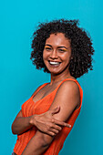 Side view of smiling African American female model in casual clothes with dark curly hair and crossed arm looking at camera against blue background