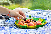 Nahaufnahme einer Hand, die eine reife Aprikose aus einem Korb auf einer Picknickdecke auswählt, vor einem natürlichen, sonnigen Hintergrund