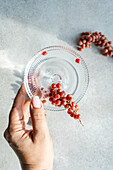 Eine zarte Hand präsentiert eine transparente Untertasse, die mit kleinen roten Beeren gefüllt ist und einen Schatten auf eine strukturierte Oberfläche wirft