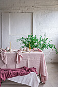 Wooden table with pink tablecloth and dishware placed near bench covered with cloth against potted plant and white brick wall