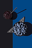 Blaubeeren in einer Schale mit Blaubeermarmelade auf einer geometrischen Serviette von oben, vor einem blauen und schwarzen Hintergrund, der einen dynamischen Schatteneffekt erzeugt