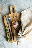 From above rustic kitchen setup on wooden cutting board with a fork, knife, a sprinkle of pink Himalayan salt, fresh rosemary sprigs, and a linen napkin, laid out on a textured concrete background
