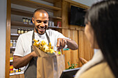 Positive young African American male seller in apron smiling and giving paper bag with products to anonymous female customer in modern supermarket